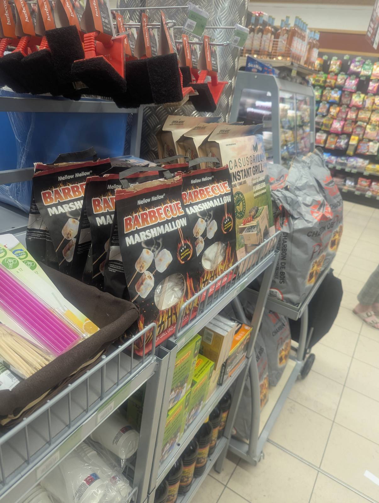 Store aisle with barbecue supplies: marshmallows, instant grill kits, brushes, and charcoal bags. Brightly colored packaging, tiled floor. The packaging for the marshmallows screams: BBQ SEASON